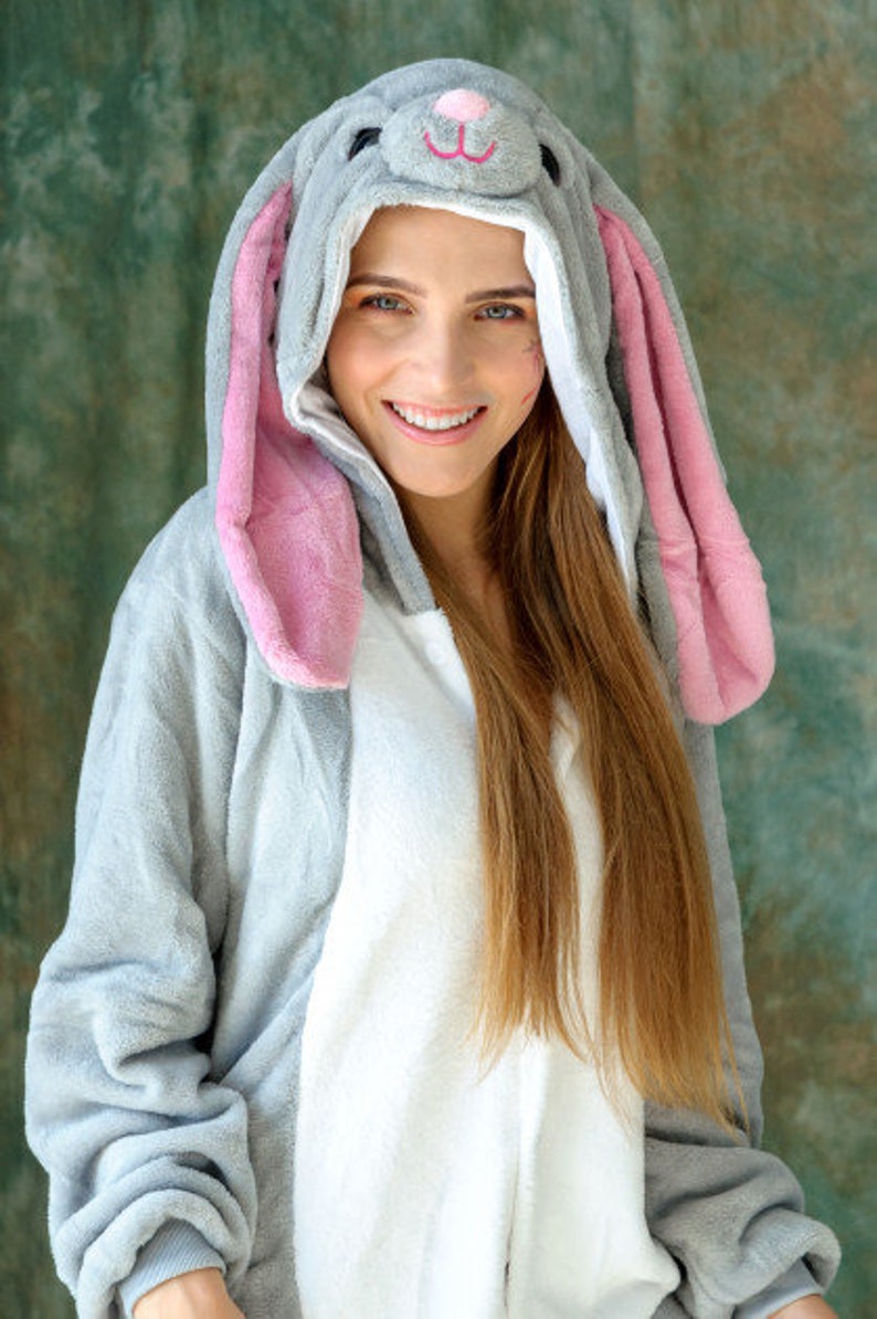 Op de afbeelding: Een vrouw draagt een grijze en roze bunny onesie met een witte voering. De onesie heeft een capuchon met bunny oren en een schattig gezicht.