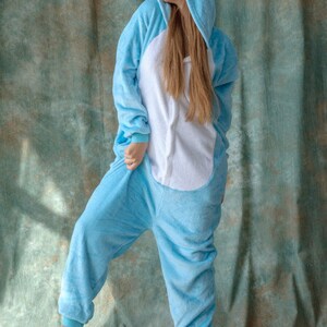May include: A person wearing a light blue hooded onesie with a white belly and bunny ears. The onesie is made of soft, plush fabric.