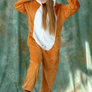 May include: A brown and white fox onesie with a hood featuring a fox face. The onesie is made of soft, plush fabric and has a zipper closure in the front.
