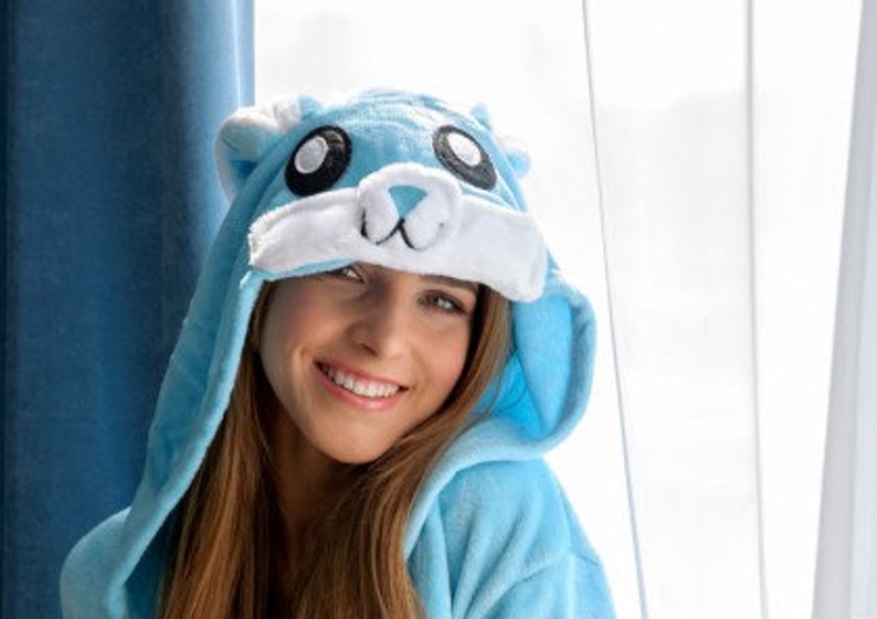 May include: A woman wearing a blue hooded onesie with a white and blue animal face on the hood. The onesie has a soft, plush texture.