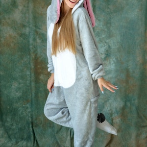 Op de afbeelding: Een grijze bunny onesie met een witte buik en roze oren. De onesie heeft een capuchon met bunny oren en een gezicht.