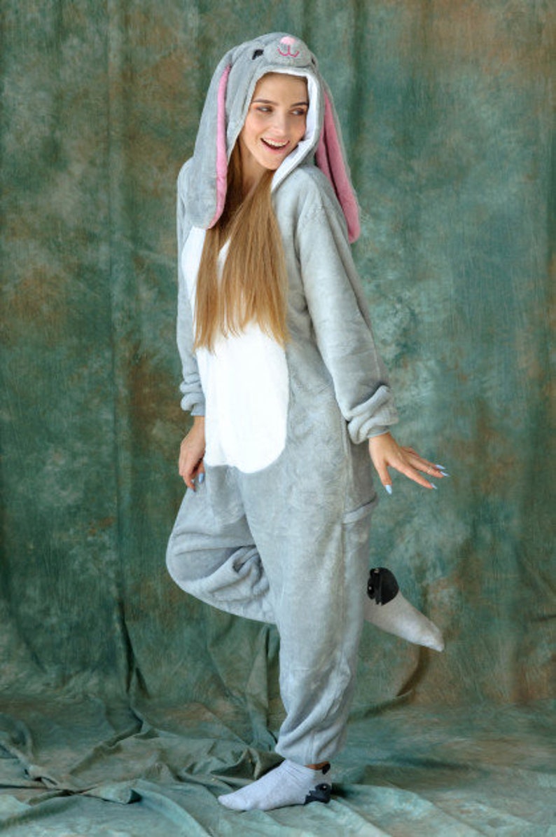 Op de afbeelding: Een grijze bunny onesie met een witte buik en roze oren. De onesie heeft een capuchon met bunny oren en een gezicht.