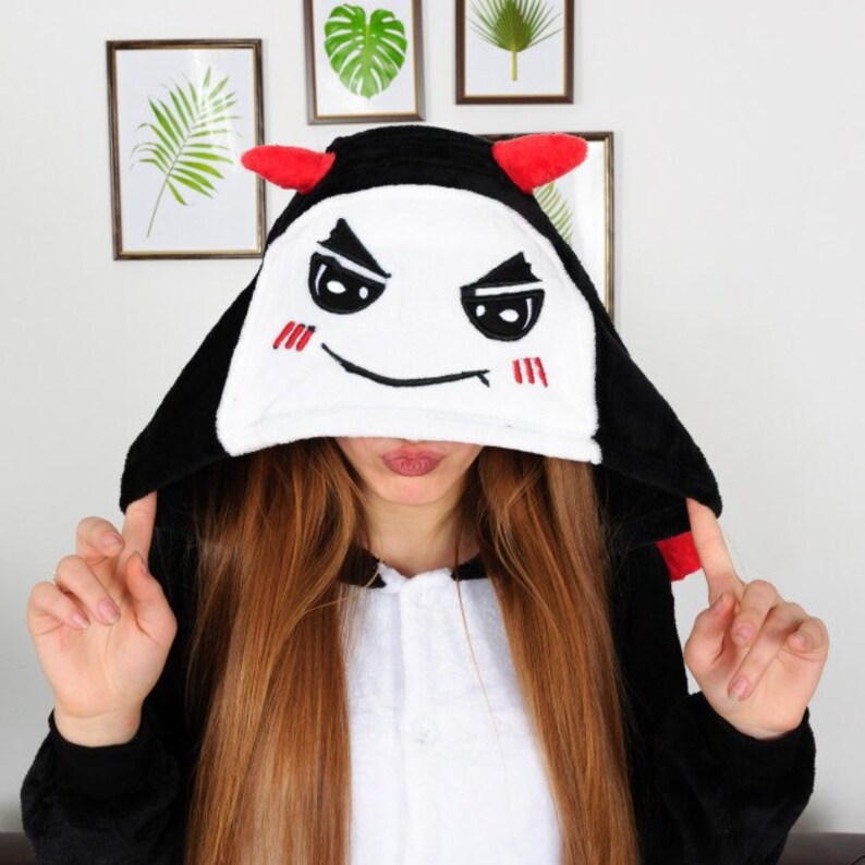 Op de afbeelding: Zwart-witte hooded onesie met een rood duivels gezicht op de capuchon. Het gezicht heeft een ondeugende grijns en rode hoorns.