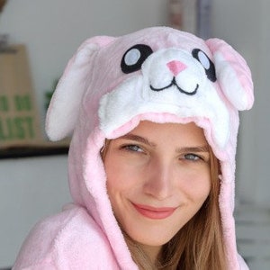 May include: A pink plush bunny hooded robe with white ears and a black nose. The hood has large black eyes and a white mouth.