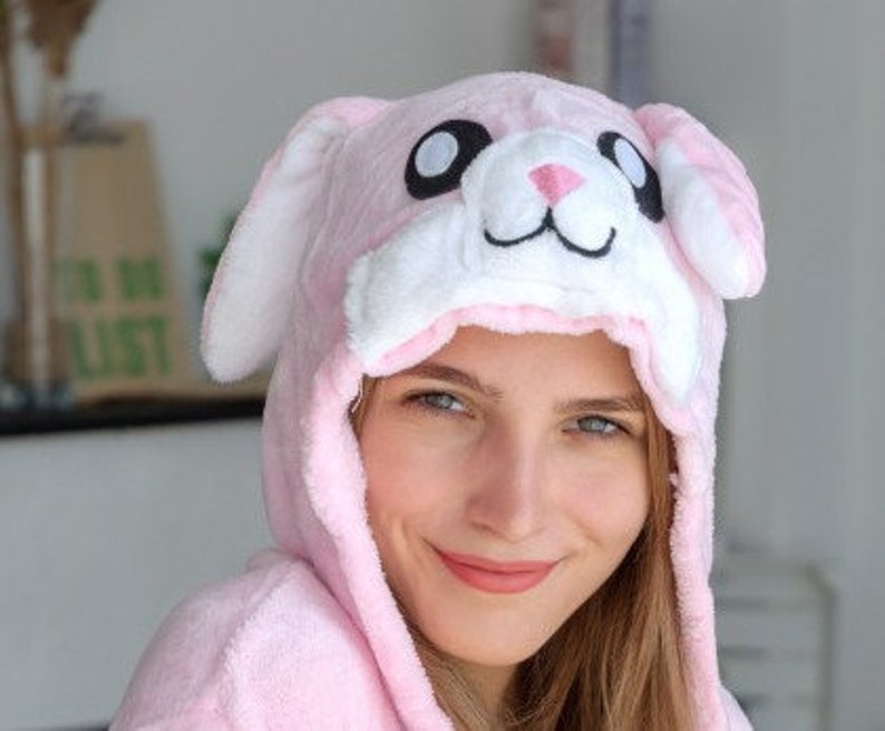 May include: A pink plush bunny hooded robe with white ears and a black nose. The hood has large black eyes and a white mouth.