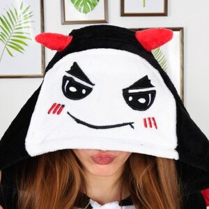 Op de afbeelding: Zwart-witte hooded onesie met een rood duivels gezicht en hoorns. Het gezicht heeft een ondeugende uitdrukking met zwarte ogen en een rode mond.