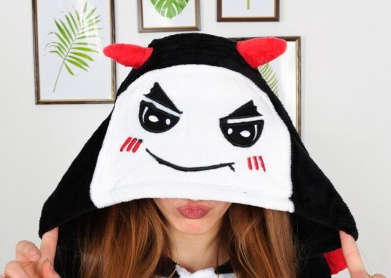 Op de afbeelding: Zwart-witte hooded onesie met een rood duivels gezicht en hoorns. Het gezicht heeft een ondeugende uitdrukking met zwarte ogen en een rode mond.