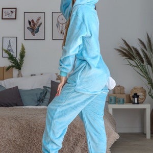 May include: A light blue fluffy onesie with a white bunny hood and tail. The onesie has a zip fastening and long sleeves. The model is wearing the onesie and is standing on a light brown rug.