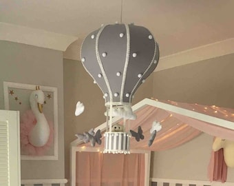 May include: A white crib with a pink canopy and a gray hot air balloon mobile hanging above it. The crib has a white mattress and a pink and white striped bedding set. There is a white wicker basket with colorful pom-poms in the corner of the room. A pink and gray round rug with a white letter K in the center is on the floor.