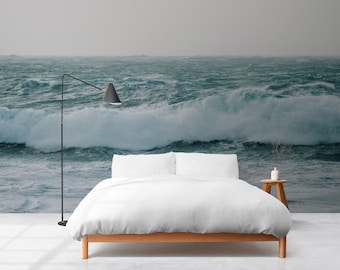 Puede incluir: Una cama blanca con un marco de madera se encuentra frente a un mural de pared que representa un océano tormentoso con grandes olas rompiendo. Una lámpara de pie negra se encuentra a la izquierda de la cama.