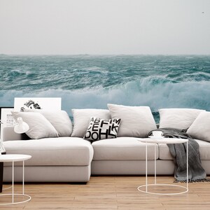 May include: A white sectional sofa with gray throw blankets and pillows. The sofa is in front of a wall mural depicting a stormy ocean with large waves.