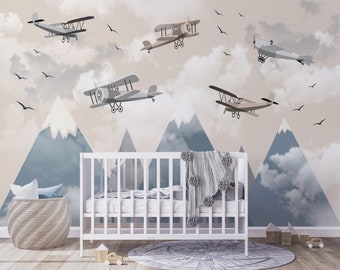 May include: A white crib with a gray pom-pom blanket in a nursery with a mountain and airplane themed mural. The mural features blue mountains, white clouds, and brown airplanes.