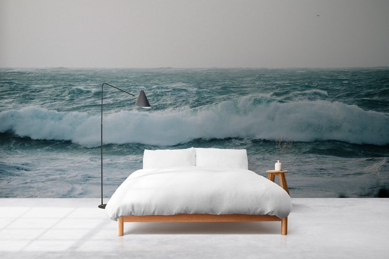May include: A white bed with a wooden frame sits in front of a wall mural depicting a stormy ocean with large waves crashing. A black floor lamp stands to the left of the bed.