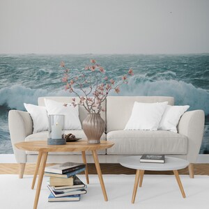 May include: A white couch with two pillows sits in front of a wall mural depicting a stormy sea. A wooden coffee table with a stack of books sits in front of the couch. A vase with pink flowers sits on the table.
