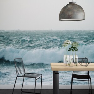 May include: A dining room with a large ocean wave mural on the wall. The room features a wooden table with black metal legs, two black metal chairs, and a rustic metal pendant light hanging above the table. There are three clear glass vases with white flowers on the table.