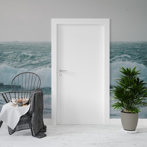 May include: A black metal chair with a gray knitted throw blanket draped over the back. The chair is in front of a white door and a stormy ocean scene wallpaper. A small white table with a bowl of fruit and a plate of cookies sits in front of the chair. A potted green plant with a black and white patterned pot sits on the floor to the right of the chair.