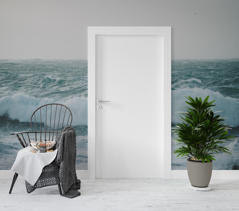 May include: A black metal chair with a gray knitted throw blanket draped over the back. The chair is in front of a white door and a stormy ocean scene wallpaper. A small white table with a bowl of fruit and a plate of cookies sits in front of the chair. A potted green plant with a black and white patterned pot sits on the floor to the right of the chair.