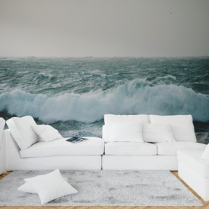 May include: A white sectional sofa with white pillows sits in front of a wall mural depicting a stormy ocean with large waves crashing.