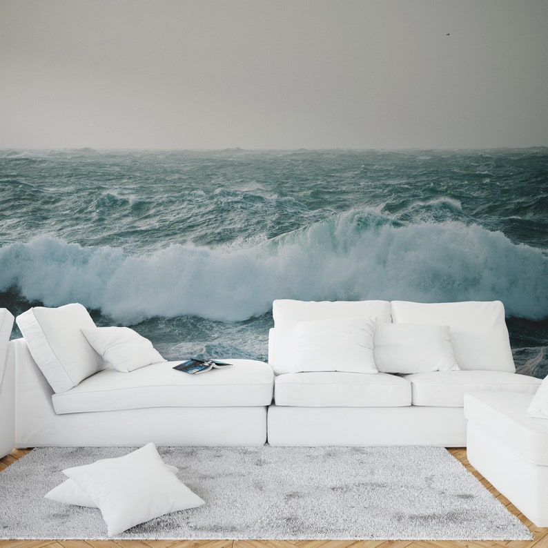 May include: A white sectional sofa with white pillows sits in front of a wall mural depicting a stormy ocean with large waves crashing.