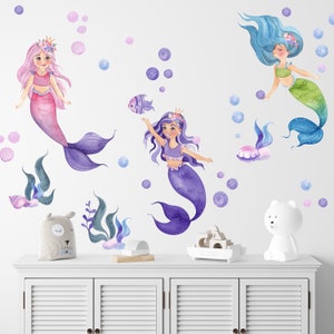May include: Three mermaid wall decals with purple, pink, and blue tails. The mermaids are wearing crowns and have long hair. The decals are surrounded by bubbles.