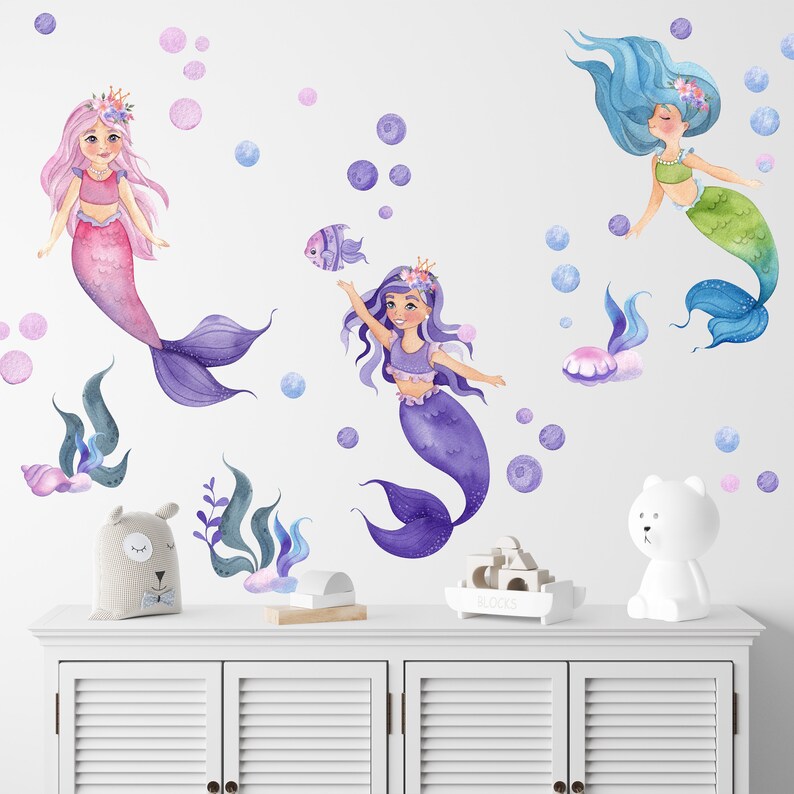 May include: Three mermaid wall decals with purple, pink, and blue tails. The mermaids are wearing crowns and have long hair. The decals are surrounded by bubbles.