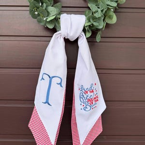 May include: White fabric scarf with red gingham trim and blue embroidered letter "T". The scarf has the phrase "Land of the Free Because of the Brave" embroidered in red and blue. A green wreath is in the background.