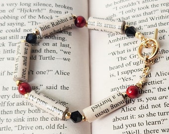 May include: A bracelet made with rolled-up pages of a book, red and black beads, and a gold chain with a clasp. The bracelet is lying on a page of a book with text about the Mock Turtle.