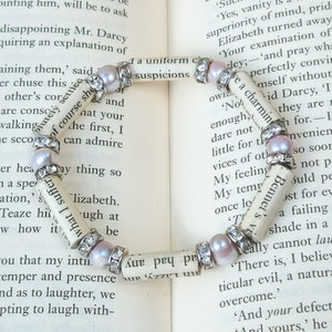 May include: A white paper bracelet with pink pearls and silver accents. The bracelet is made from pages of a book with the text "We a charming" visible. The bracelet is lying on a book.