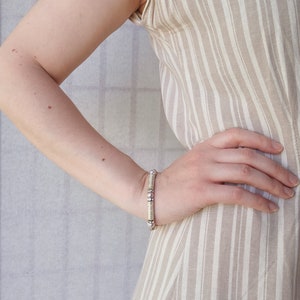 May include: A close-up of a person's arm wearing a white and silver beaded bracelet. The bracelet is made of small, round beads that are strung together on a silver chain. The person is wearing a striped dress.