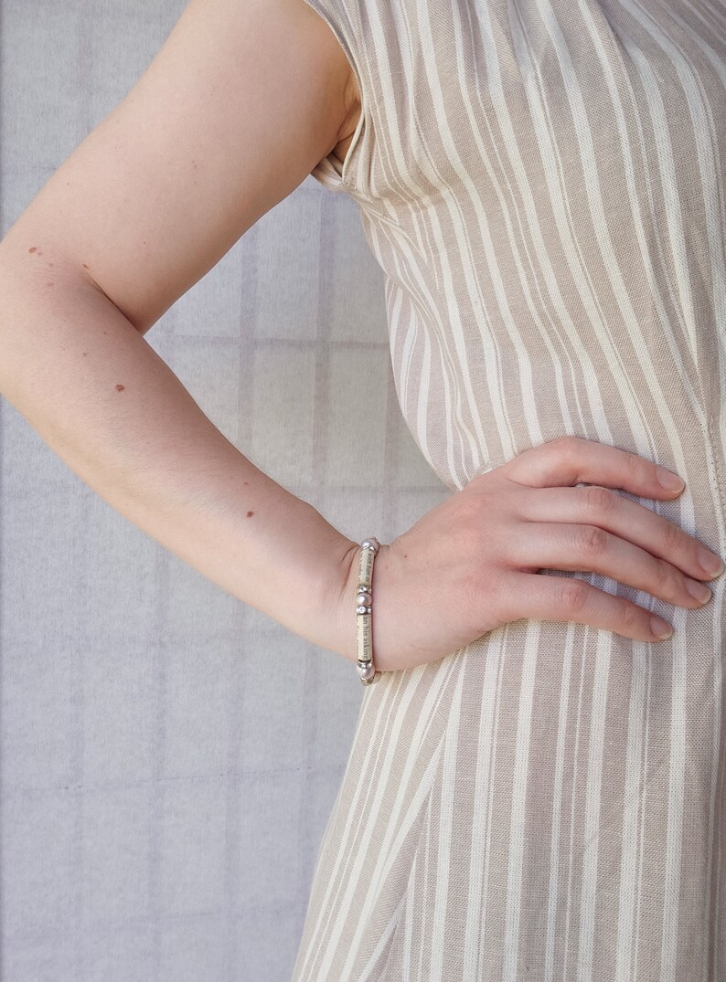 May include: A close-up of a person's arm wearing a white and silver beaded bracelet. The bracelet is made of small, round beads that are strung together on a silver chain. The person is wearing a striped dress.