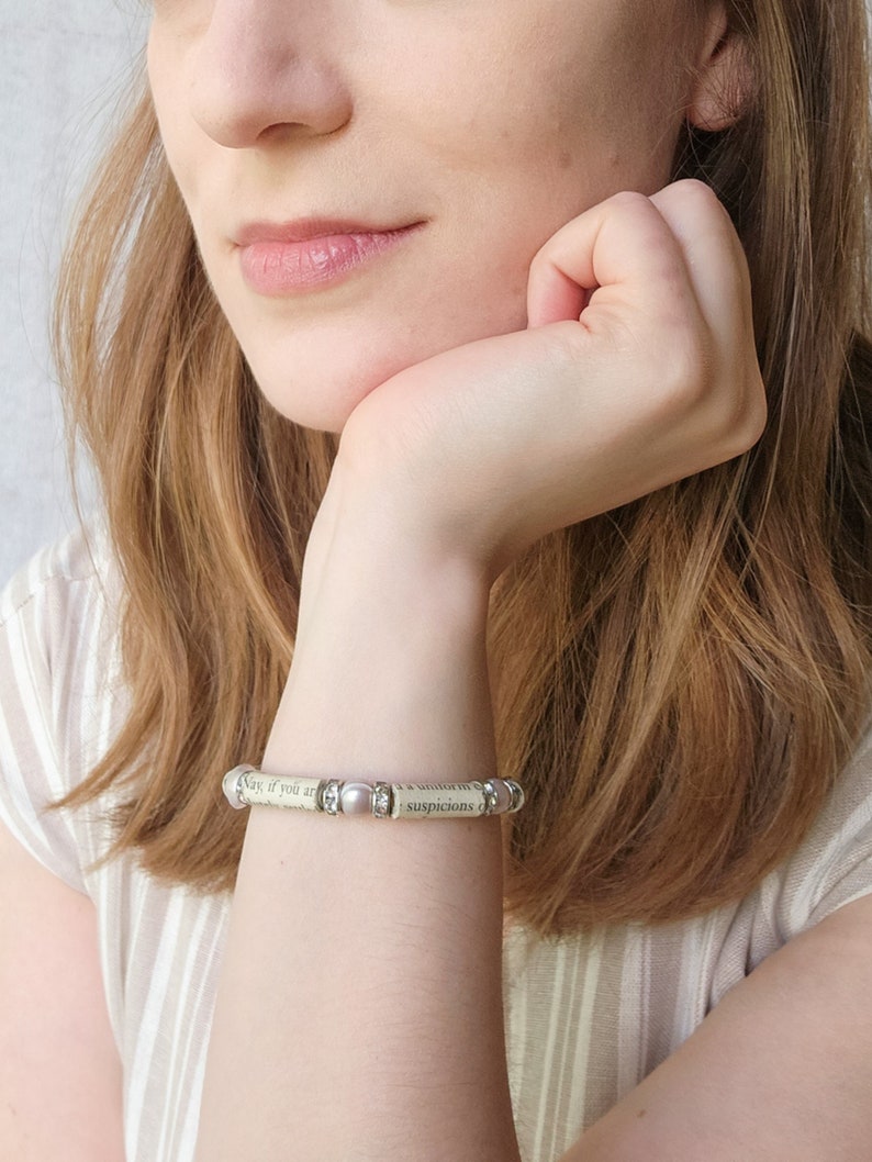 May include: A bracelet made of a book page with small pearl-like beads and silver accents. The bracelet reads "Nay, if you are a villain in the matter, you must needs be a villain of the first magnitude, and I have a villainous suspicion that you are so."