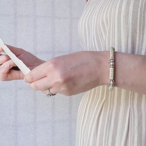 May include: A close-up of a person's wrist wearing a bracelet made of paper with pink beads. The bracelet has text printed on it.