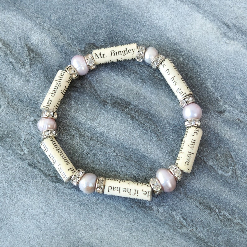 May include: A bracelet made of small, pale pink beads and paper scrolls with text from the book "Pride and Prejudice" printed on them. The text includes the phrases "Mr. Bingley", "her daughter", "to his gall", "te, my love", "if he had", "Pride obser", "sometimes", "state, but did".