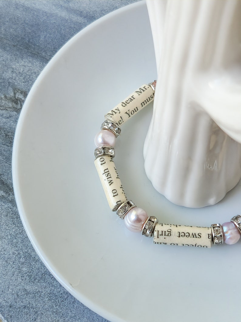May include: A bracelet made of small white beads with pink accents and silver accents. The beads are made of paper with text printed on them. The text reads "My dear Mr. ... a sweet girl. ... to wish to ... with her h ... e! You must".