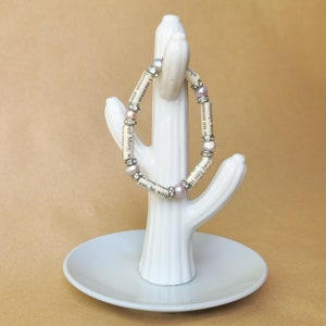 May include: A white ceramic cactus ring holder with a pink and silver beaded bracelet wrapped around it. The bracelet is made of book pages and has small pink beads.