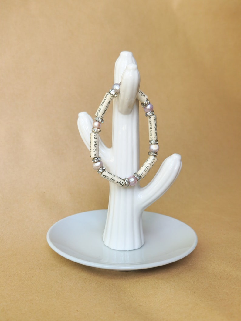May include: A white ceramic cactus ring holder with a pink and silver beaded bracelet wrapped around it. The bracelet is made of book pages and has small pink beads.