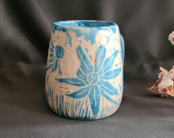 May include: A handmade ceramic mug with a cream-colored base and blue floral design. The mug features a cylindrical shape with a handle and is decorated with carved blue flowers and foliage. The mug is approximately 4 inches tall.
