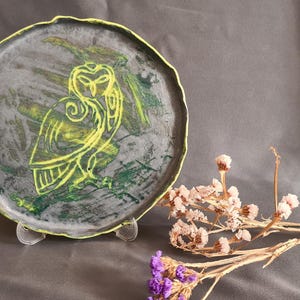May include: A handmade ceramic plate with a gray background and a yellow owl design. The plate has a wavy edge with a yellow border. Dried flowers in shades of pink and purple are in the foreground.