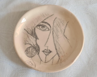 May include: A white ceramic plate with a black and white line drawing of a woman's face. The woman's eye is visible, and her hair is drawn in a stylized way.