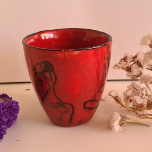 Peut inclure: Une petite tasse en céramique rouge avec des motifs abstraits noirs. La tasse est émaillée et a une finition brillante.