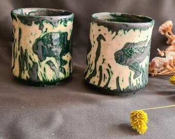 May include: Two handmade ceramic cups with a cream-colored base and green and black accents. Each cup features a turtle design. The cups are cylindrical with a slightly irregular shape. Dried flowers and a yellow flower are in the background.