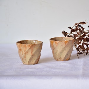 Könnte beinhalten: Zwei handgefertigte Keramikbecher mit einem einzigartigen, strukturierten Design. Die Becher haben eine beige Grundfarbe mit braunen und orangefarbenen Akzenten und ein gesprenkeltes Finish. Die Becher werden auf einer weißen Oberfläche präsentiert.