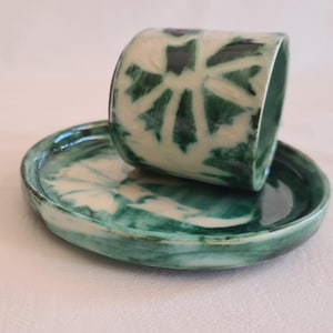 Può includere: Un set di tazza e piattino in ceramica. La tazza è cilindrica con un motivo floreale verde e bianco. Il piattino è rotondo e verde, con un motivo floreale abbinato. Il set è fatto a mano e ha un'estetica rustica.