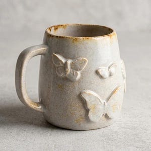 May include: A speckled light gray ceramic mug featuring raised, three-dimensional butterfly motifs on the exterior surface. The mug has a sturdy handle and a slightly uneven, textured rim with brown accents along the top edge of the vessel.