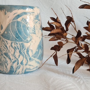 Könnte beinhalten: Eine Keramikvase mit einem blauen und weißen Design von Fischen und Seetang. Die Vase steht auf einer weißen Oberfläche mit einem braunen Zweig mit getrockneten Blättern.