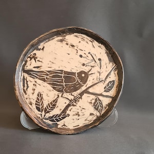 Può includere: Un piatto rotondo in ceramica con un disegno di uccello e ramo. L'uccello è marrone scuro, posato su un ramo con foglie, su uno sfondo color crema. Il bordo del piatto è marrone scuro.
