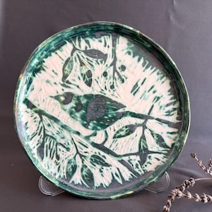 May include: A round ceramic plate featuring a sgraffito design of a bird perched on a branch amidst leaves. The surface displays a high-contrast color scheme with deep forest green glaze accents layered over a creamy white base, creating a textured look.