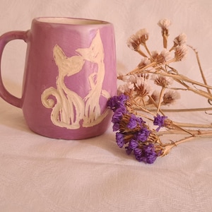 Puede incluir: Una taza de cerámica morada con una silueta blanca de dos gatos. La taza está sobre una superficie blanca con flores secas.