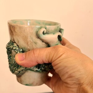 May include: A ceramic mug with a green and brown glaze. The mug has a unique design with a raised, textured, green and white floral pattern.