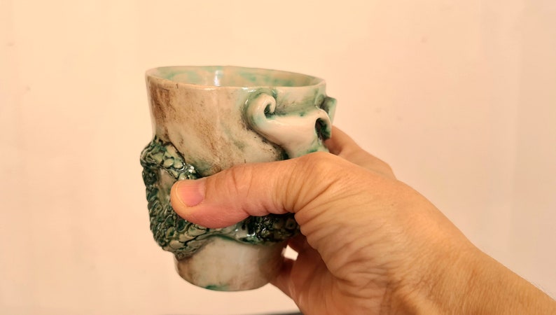 May include: A ceramic mug with a green and brown glaze. The mug has a unique design with a raised, textured, green and white floral pattern.